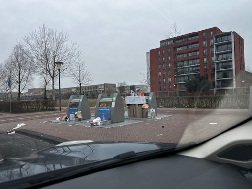 Afvalcontainers op een parkeerplaats, omringd door rommel en een gebouw op de achtergrond.