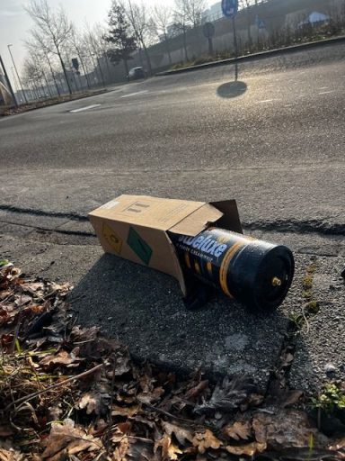 Een geopend kartonnen doos met een zwarte en gele zaklamp ligt op de grond naast een weg.