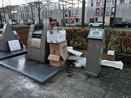 Afvalcontainers met stapels dozen en restafval ernaast in een stadsomgeving.
